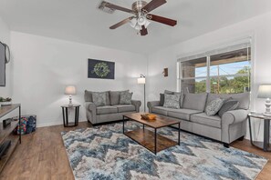 Un salon avec deux canapés gris, une table basse, un ventilateur de plafond et une grande fenêtre.