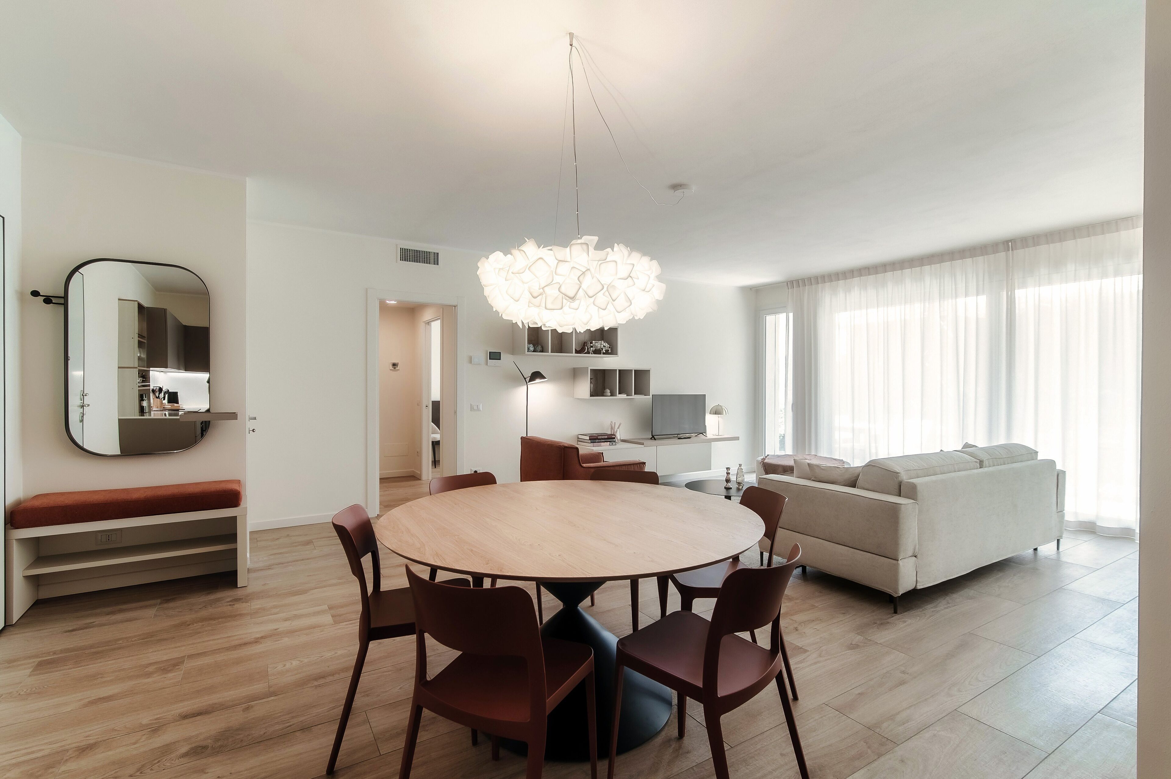 Een moderne woonkamer met een ronde houten eettafel omringd door stoelen, een bank, een televisie en een grote spiegel aan de muur.
