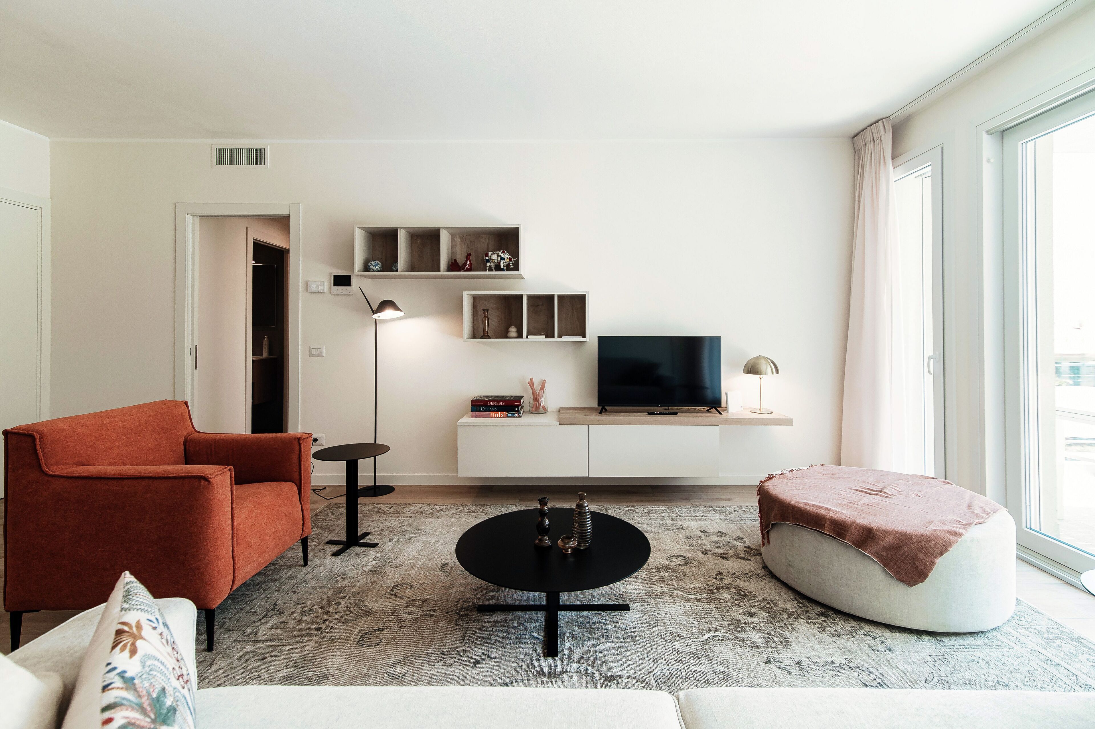 A modern living room with a sofa, ottoman, coffee table, TV, shelves, and a window.