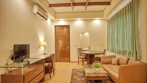 A modern living room with a glass coffee table, leather sofa, wooden TV stand, and a dining area with chairs and a table. - Enkay Suites - DLF CyberCity (Gurugram)