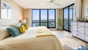 Un dormitorio con una cama grande, ventilador de techo, vista al exterior a través de una puerta corrediza de vidrio y un aparador con cajones.
