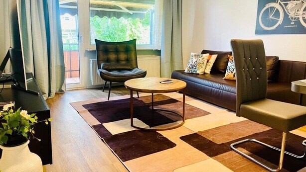 Ein modernes Wohnzimmer mit einer Couch, einem runden Couchtisch und einem Sessel. Ein Fenster mit Vorhängen, ein Heizkörper und ein Wandbild mit einem Fahrrad sind zu sehen.