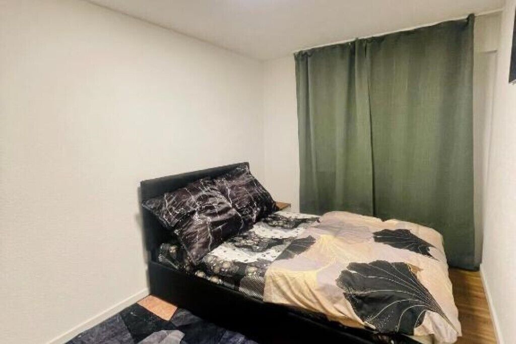 Una habitación pequeña con una cama individual, ropa de cama oscura, cortina verde y colcha estampada.