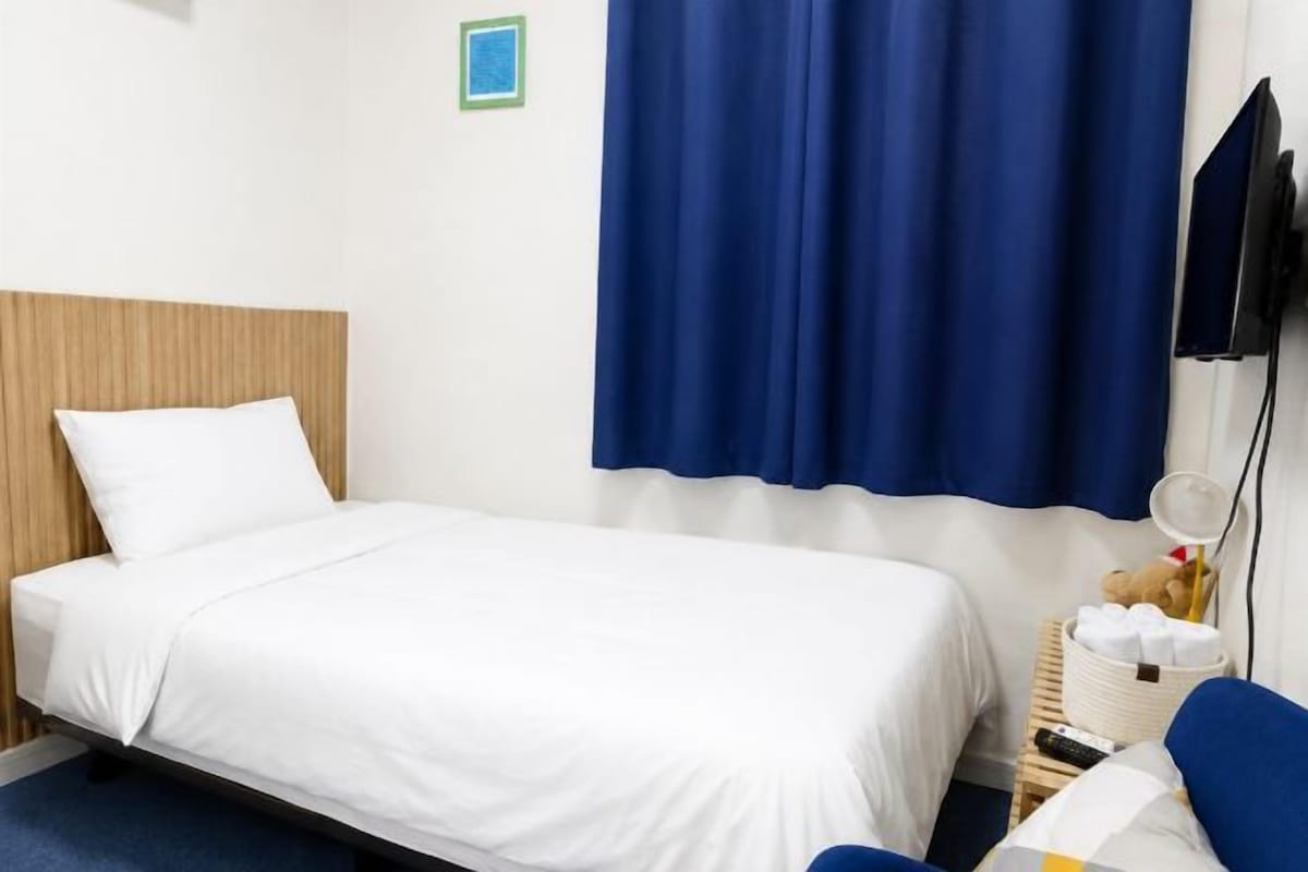 A single bed with white bedding, a wooden headboard, a blue curtain, and a small bedside table with a lamp and a teddy bear.