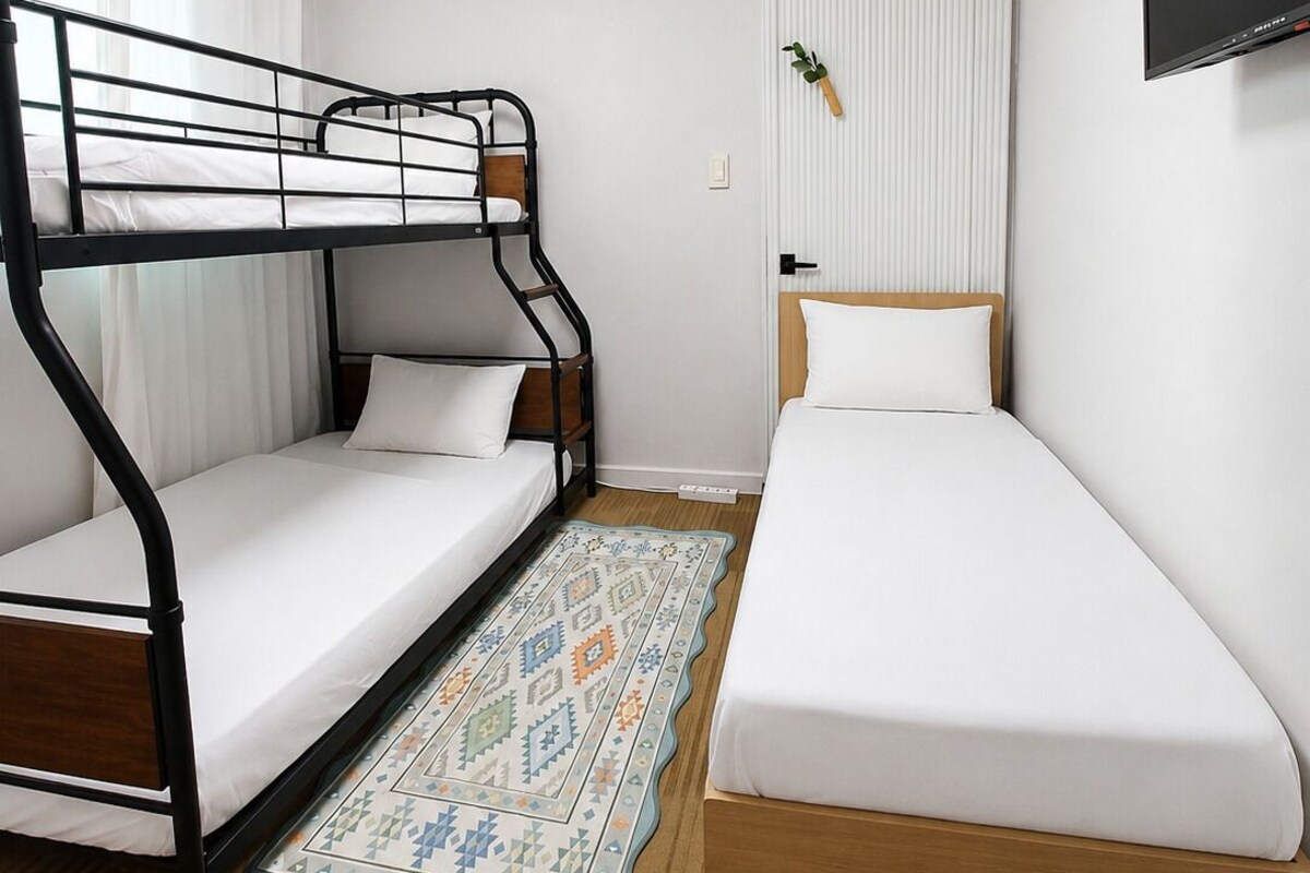 A room with a bunk bed, a single bed, a TV mounted on the wall, and a patterned rug on the floor.