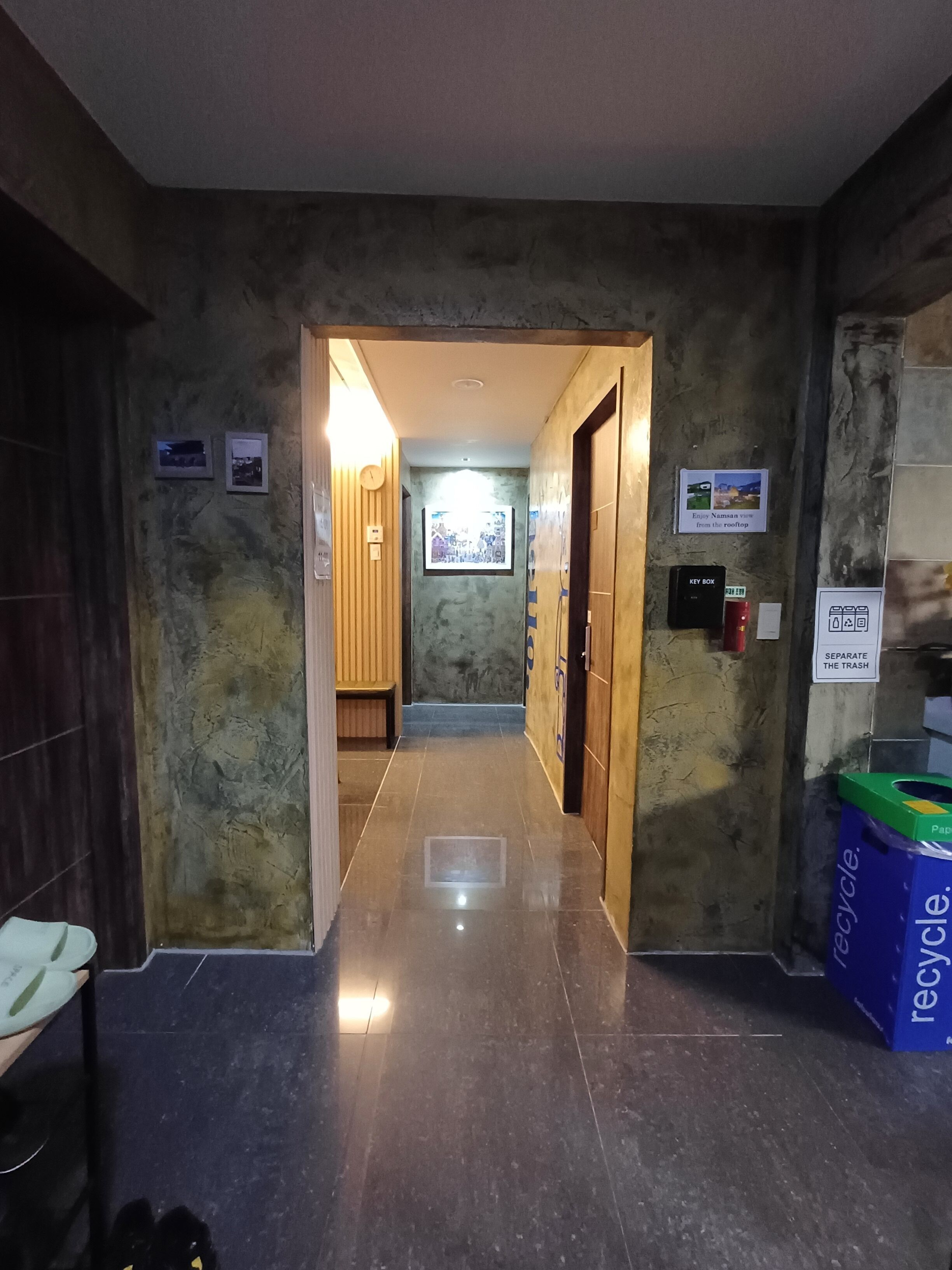 A hallway with a wooden bench, a wall-mounted clock, and a recycling bin.