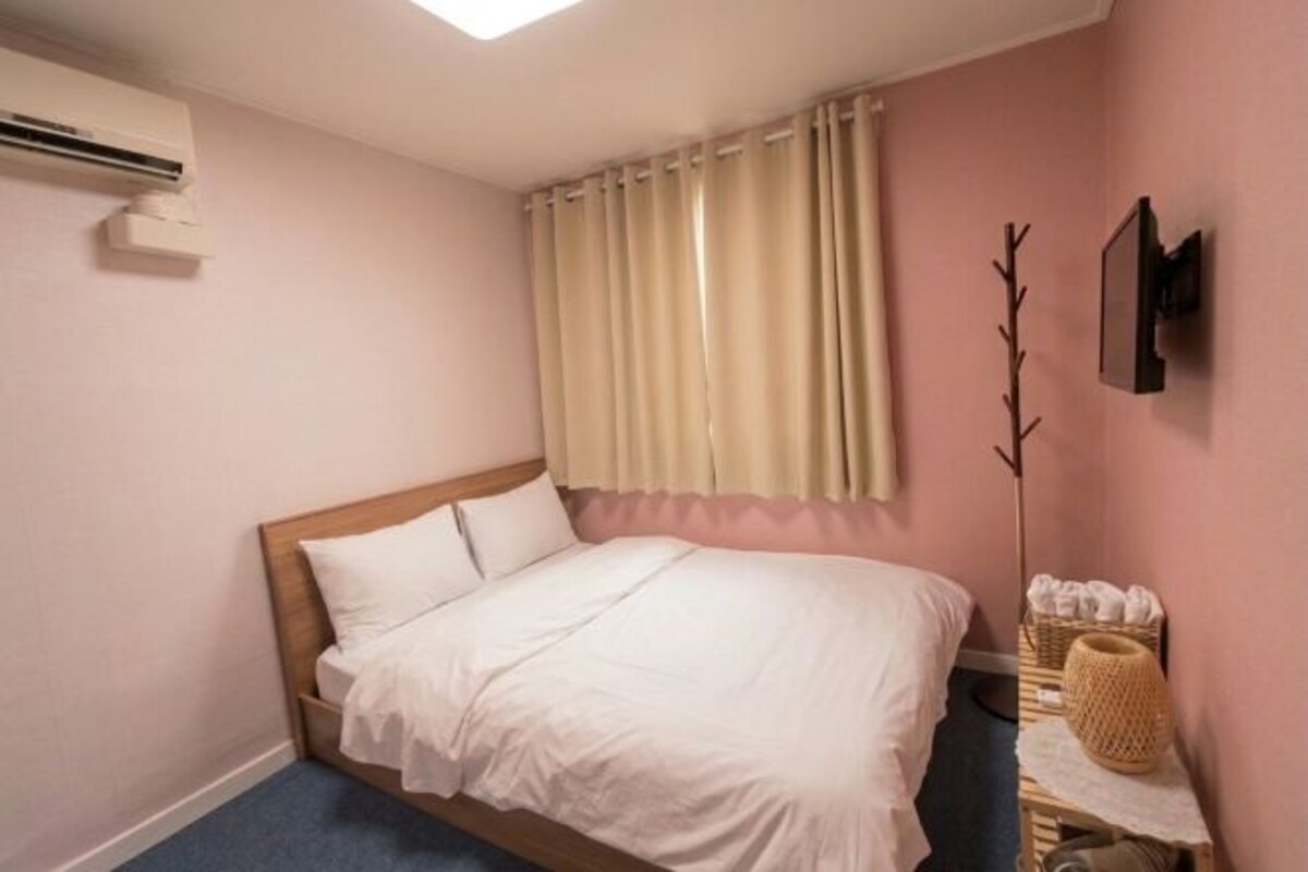 A bedroom with a bed, a window with curtains, a wall-mounted air conditioner, and a small table with a basket.