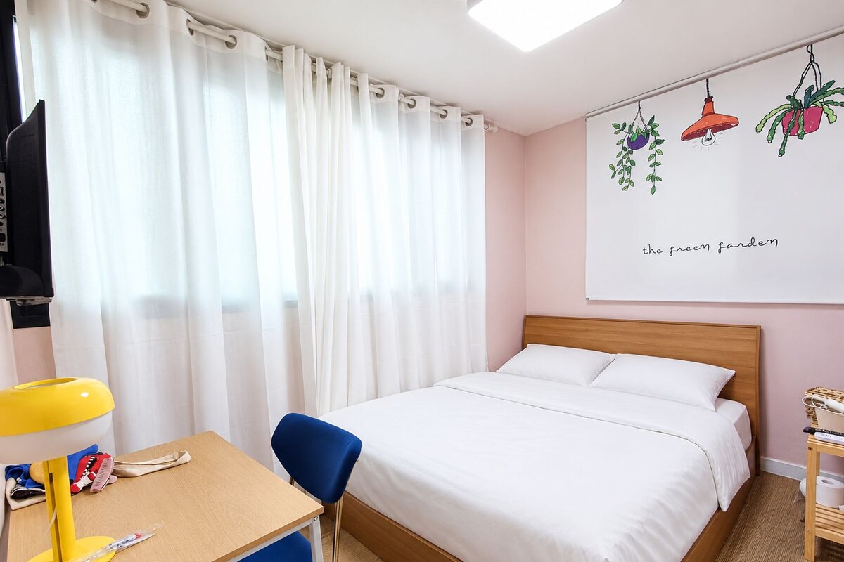 A hotel room with a bed, a desk with a chair, a lamp, and a wall-mounted artwork titled 'the green garden'.