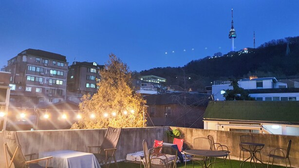A rooftop terrace with outdoor seating, a view of a cityscape including a tower, and a tree. - Space Guesthouse (Seoul)