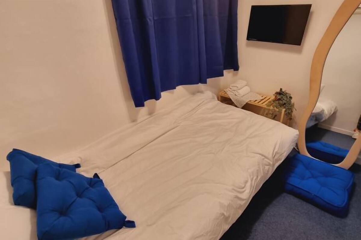 Una cama con ropa blanca, almohadas azules, una mesita con una planta, un televisor en la pared y un espejo con marco de madera.
