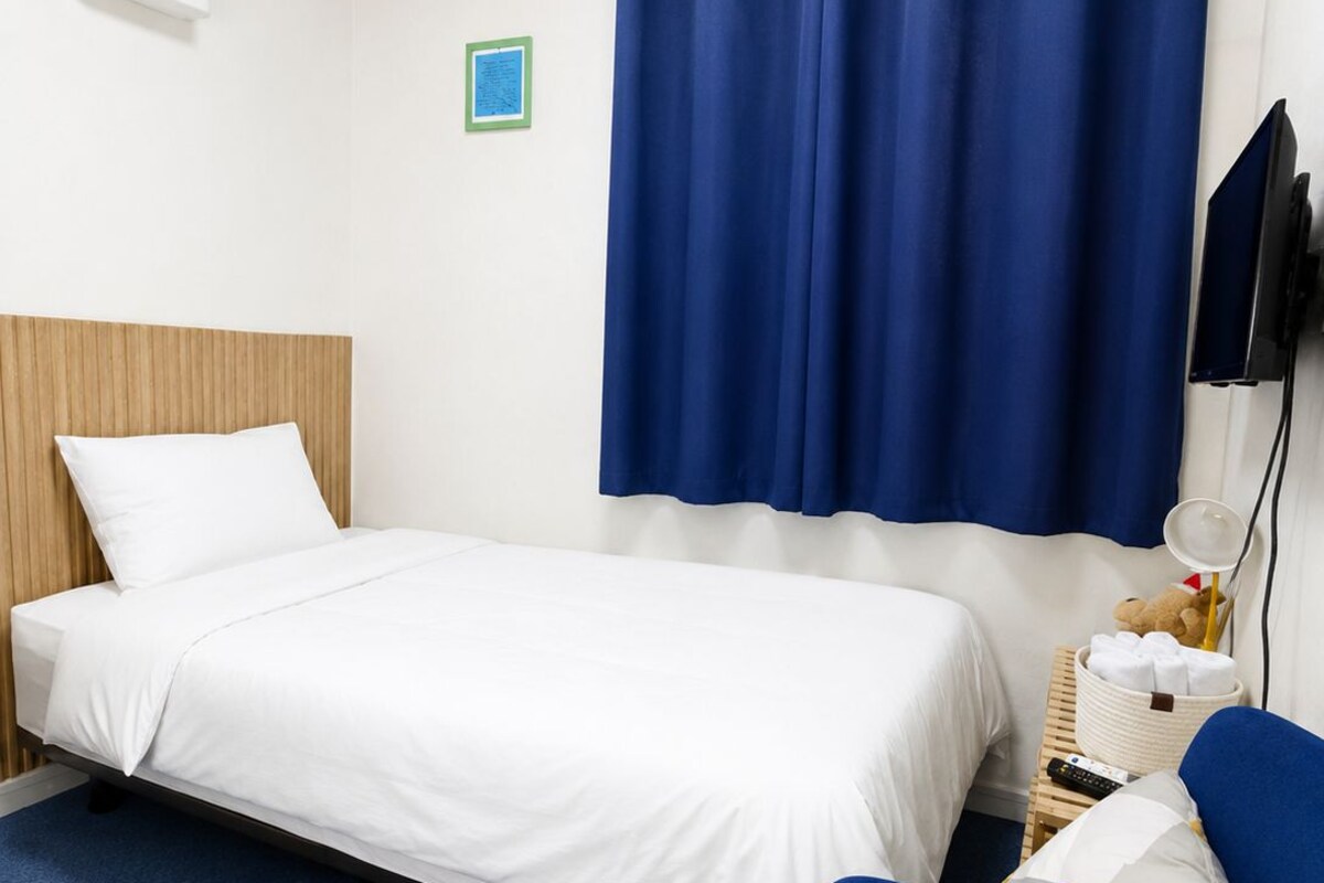 A hotel room with a bed, a chair, a television, a bedside table with a lamp, and a wall-mounted air conditioning unit.