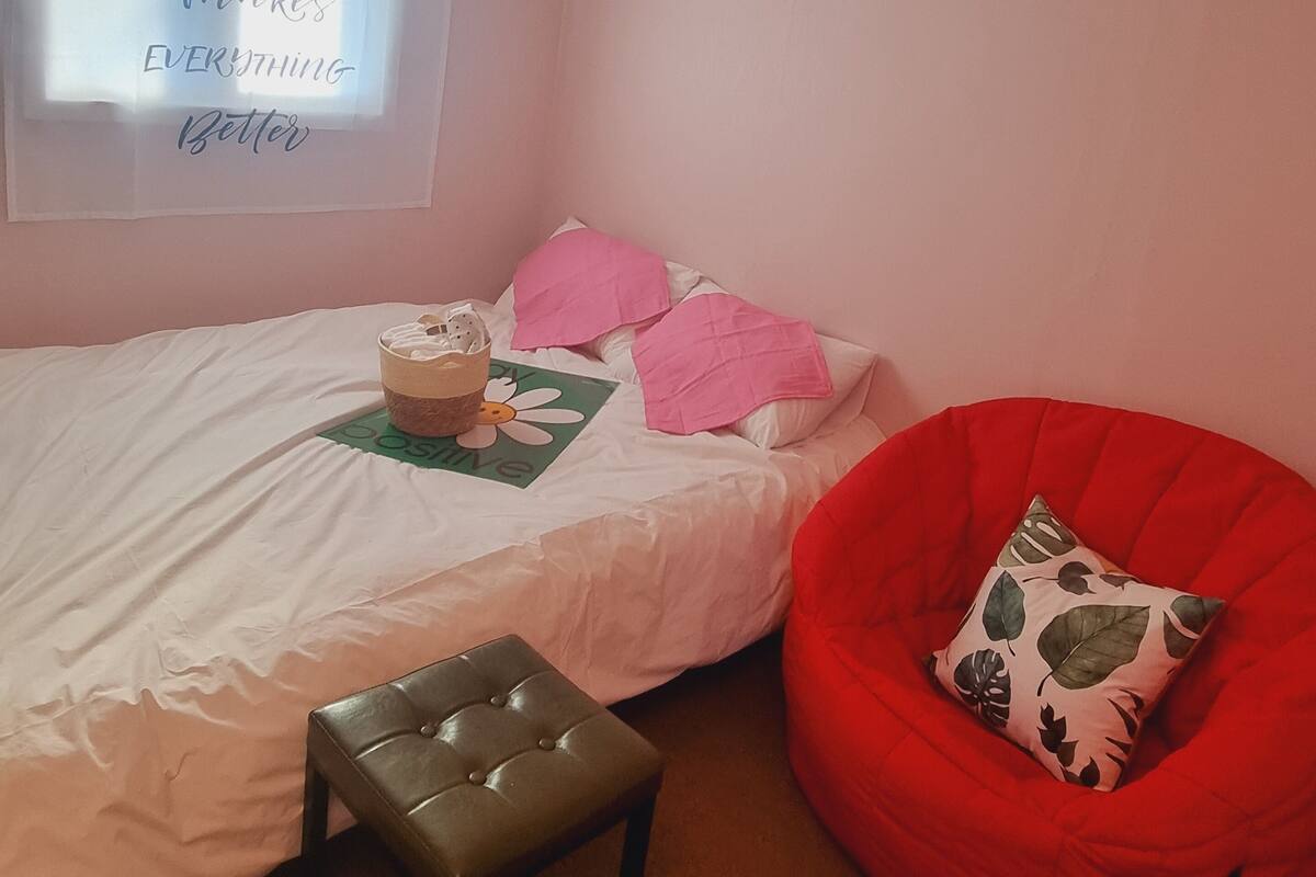 Un dormitorio con una cama, un sillón de peluche rojo, un taburete pequeño y una ventana con una frase motivacional.