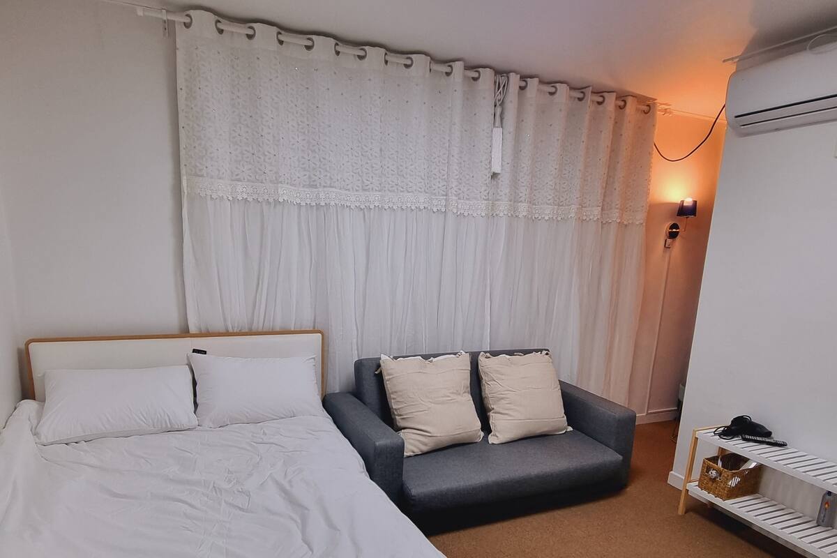 Un dormitorio con una cama, un sofá gris, una mesita con una lámpara y una ventana con cortinas.