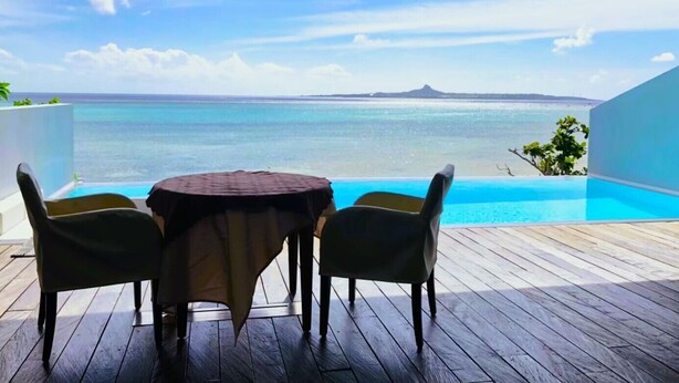 A wooden deck with a round table and chairs, overlooking a clear blue sea and a distant island. - Secret Pool Villa SEJI (Motobu)