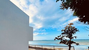 A coastal view with a white building, a tree, and a sandy beach with a fence. - Secret Pool Villa SEJI (Motobu)