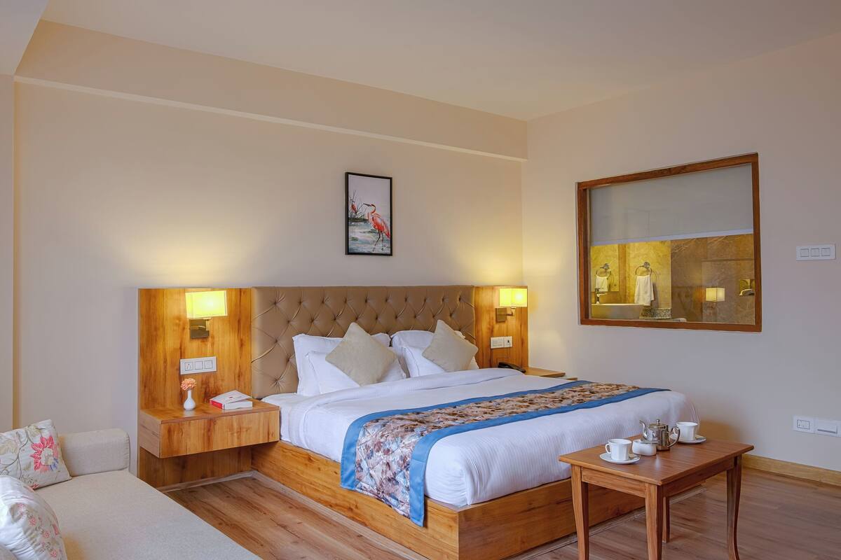 A hotel room with a large bed, wooden headboard, bedside lamps, a bedside table with a vase and book, a small table with cups and a teapot, and a painting on the wall.