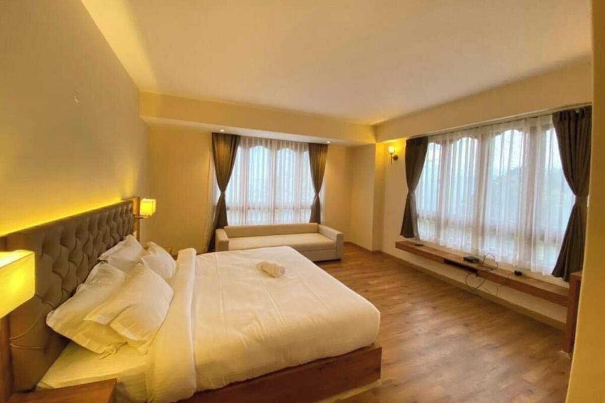 A hotel room with a large bed, a sofa, a window with curtains, and a wall-mounted lamp.