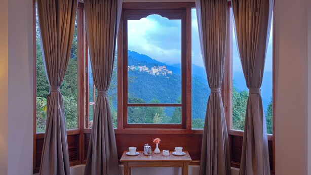 Ein Zimmer mit Holztisch, gedeckt für Tee, Vorhängen und Blick auf eine gebirgige Landschaft.
