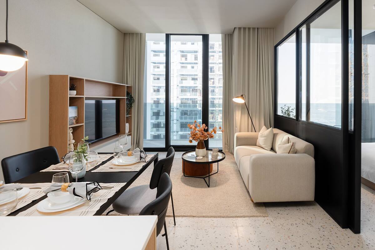 A modern hotel room with a dining area, a sofa, a bed, and large windows offering city views.
