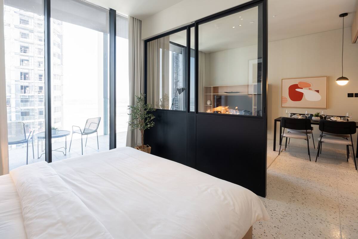 A modern hotel room with a large bed, a dining area with two chairs, a black partition, and a view of the city through large windows.
