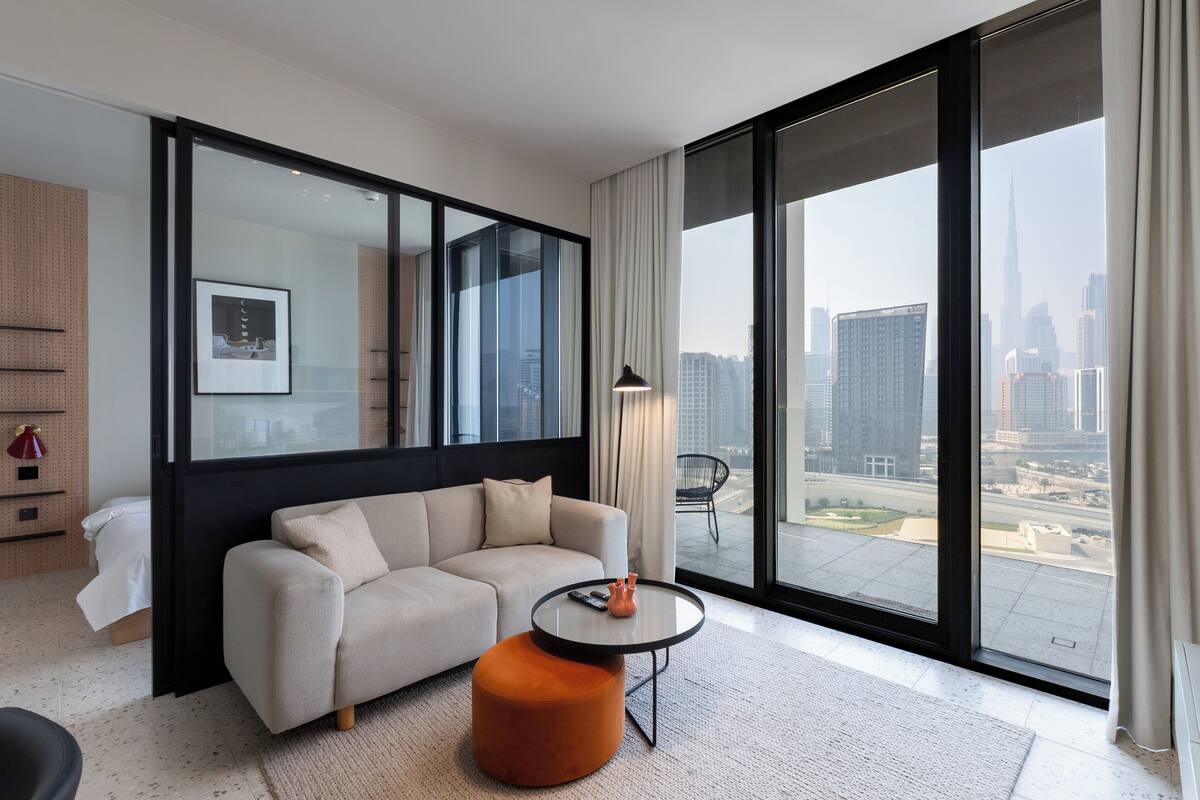 A modern hotel room with a large window, a sofa, a coffee table, and a view of the cityscape.