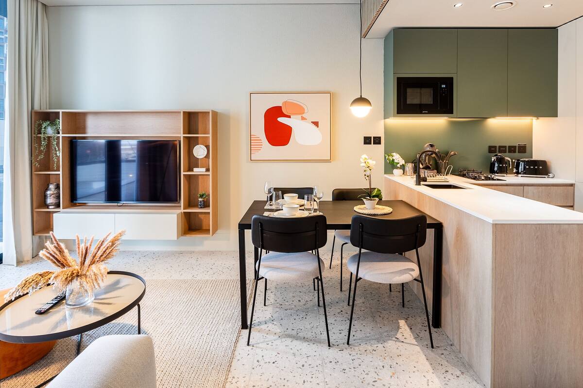 A modern living room with a TV, dining table, and chairs. There is a kitchen area with a microwave and sink.