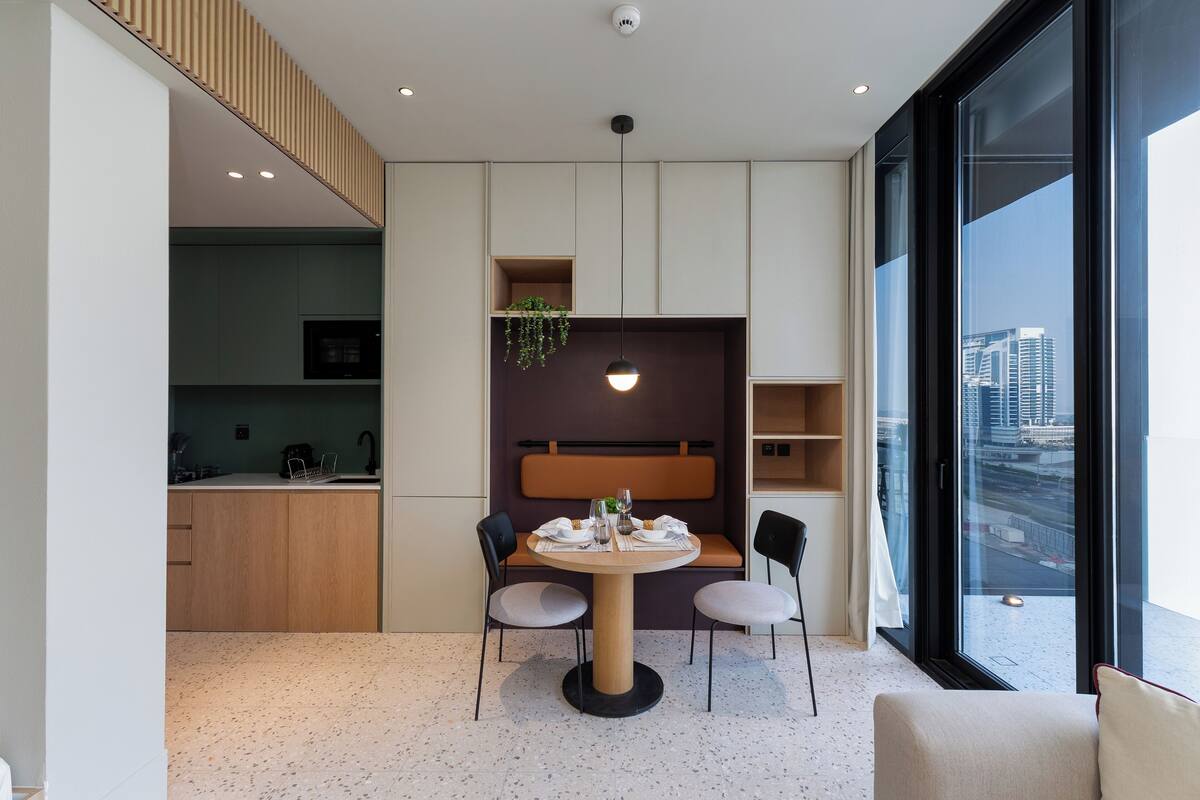 A modern dining area with a round table, two chairs, a hanging light fixture, and a view of the city through large windows.