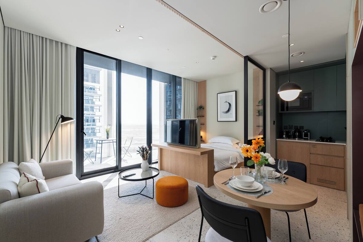 A modern hotel room with a sofa, a dining table set, a kitchen area, and large windows offering a city view.