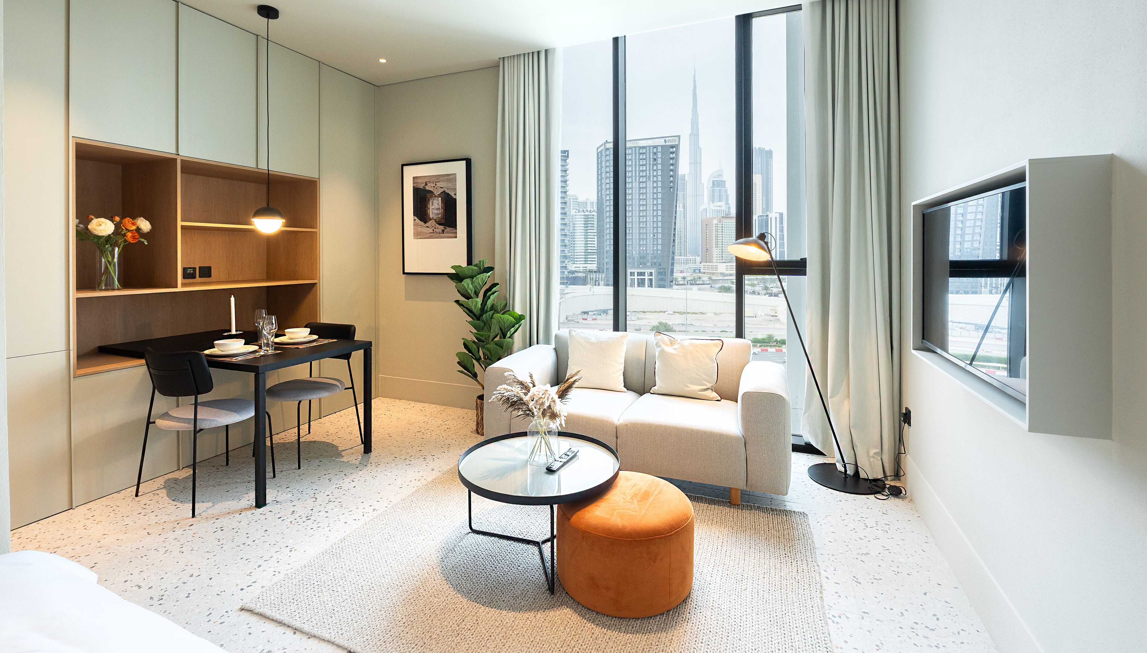 Une chambre d’hôtel moderne dotée d’un coin repas, d’un canapé, d’un pouf rond et d’une grande fenêtre donnant sur la ville.