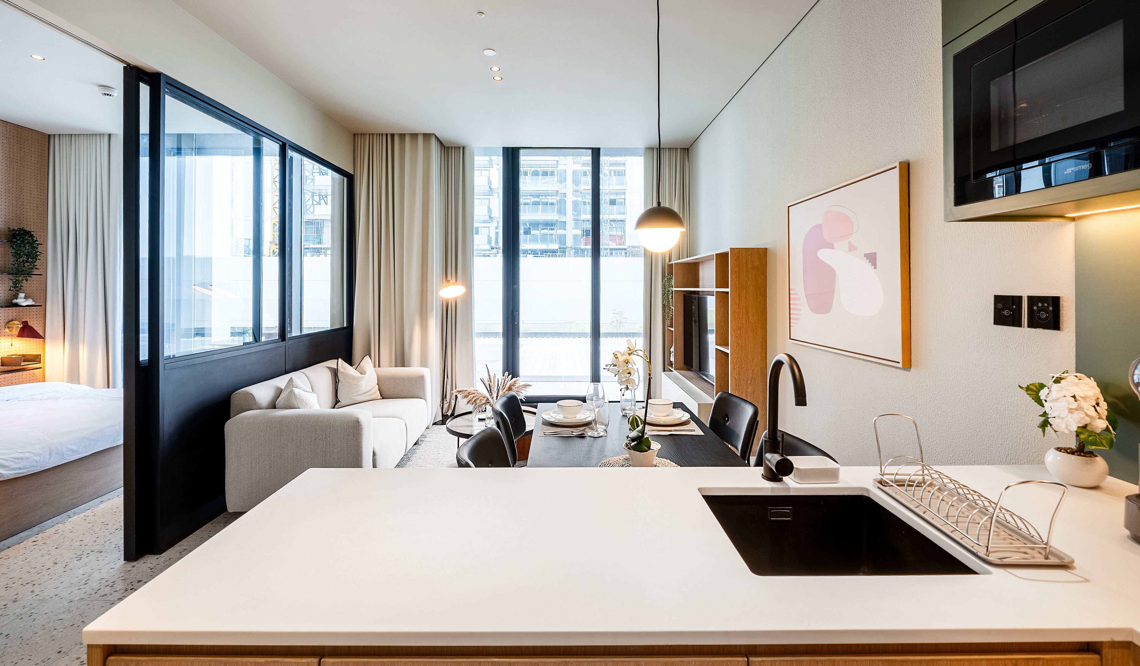 Habitació d'hotel moderna amb una finestra gran, un sofà, una taula de dinar amb cadires, una zona de cuina i un televisor de pantalla plana penjat a la paret.