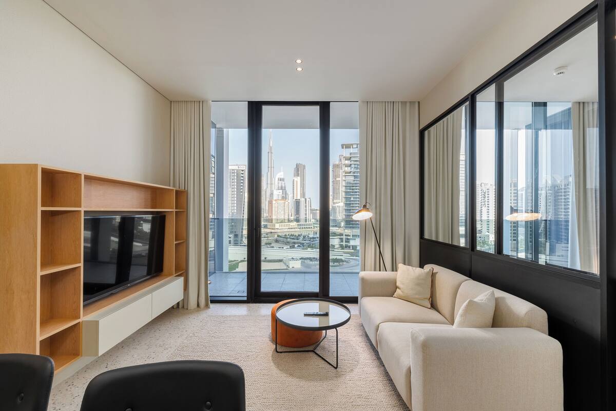 A modern living room with a large TV, a sofa, a coffee table, and a view of the city skyline through large windows.