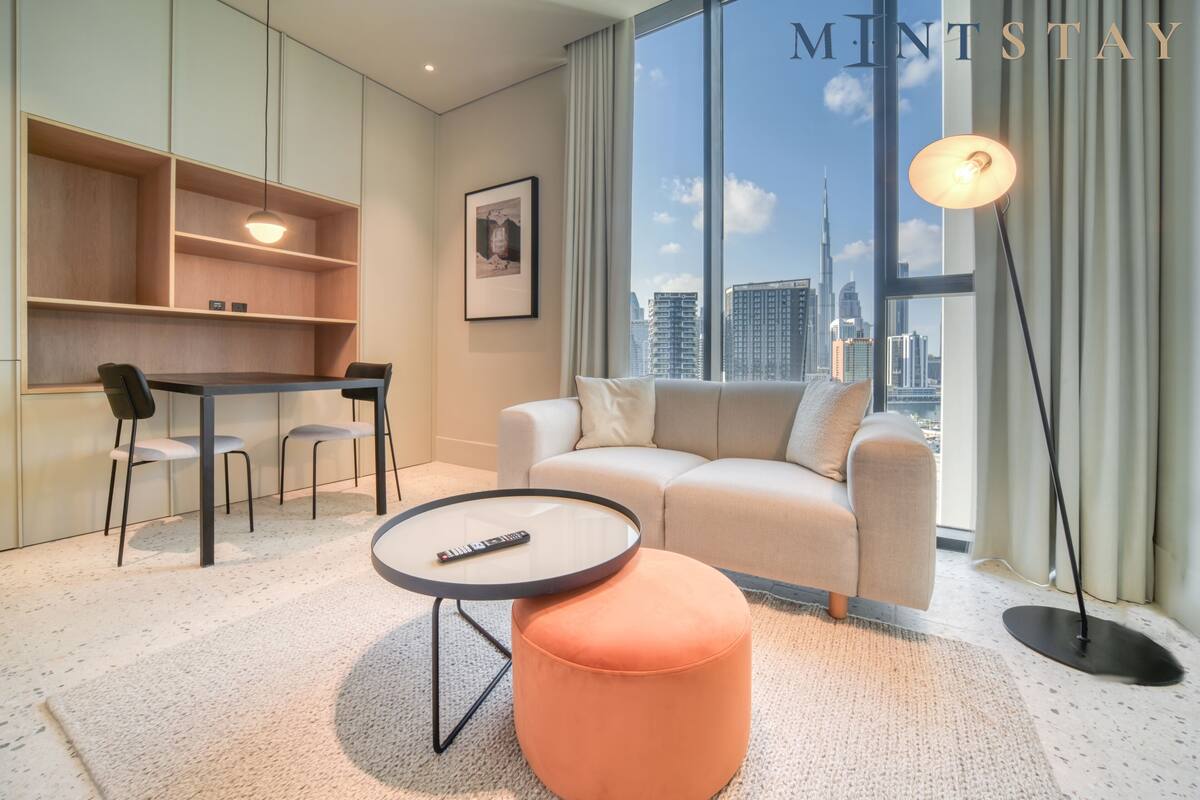 A modern living room with a sofa, ottoman, and a round coffee table. There is a desk with chairs and a bookshelf. A framed picture hangs on the wall. The room has a large window with a view of a cityscape.