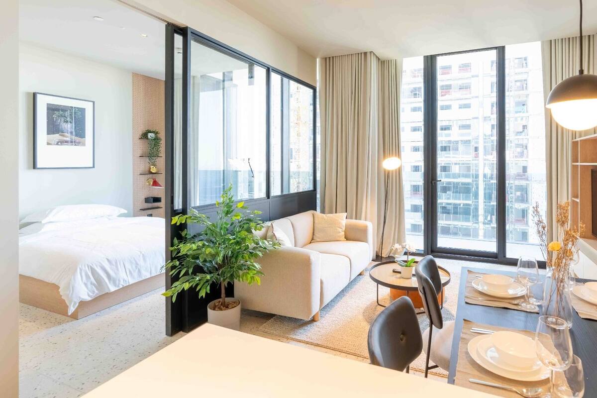 A hotel room with a bed, a sofa, a dining table set for two, and a large window with a city view.
