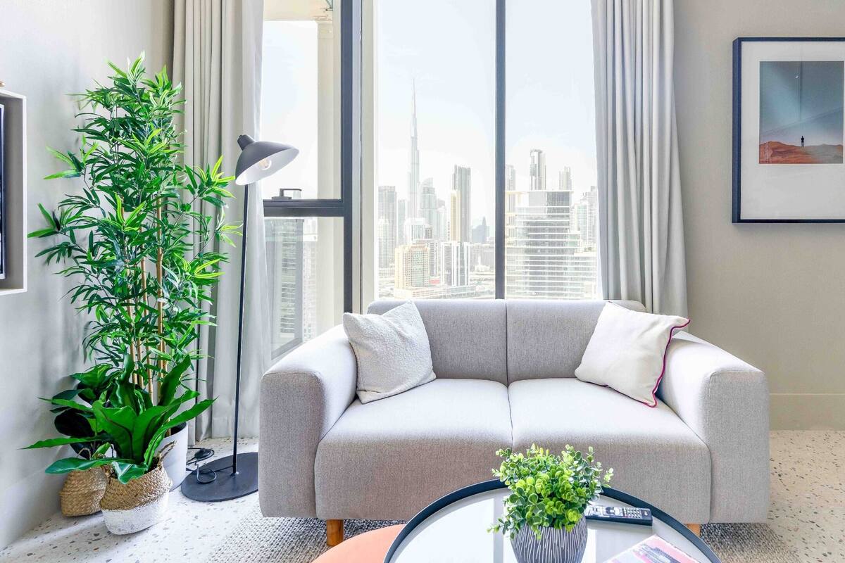 A modern living room with a grey sofa, a large potted plant, and a city view through large windows.