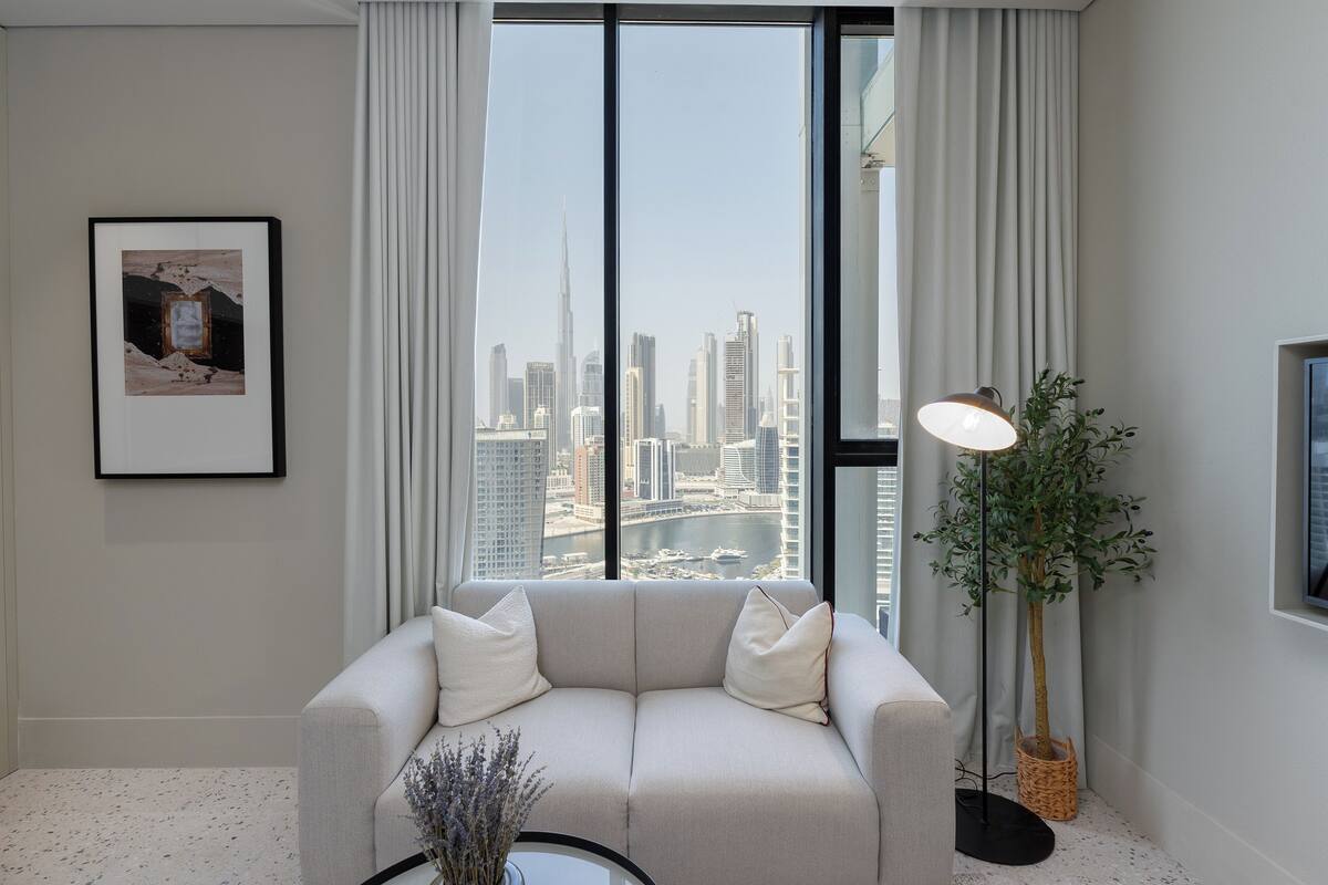 A modern living room with a large window, a sofa, a floor lamp, and a framed picture on the wall.