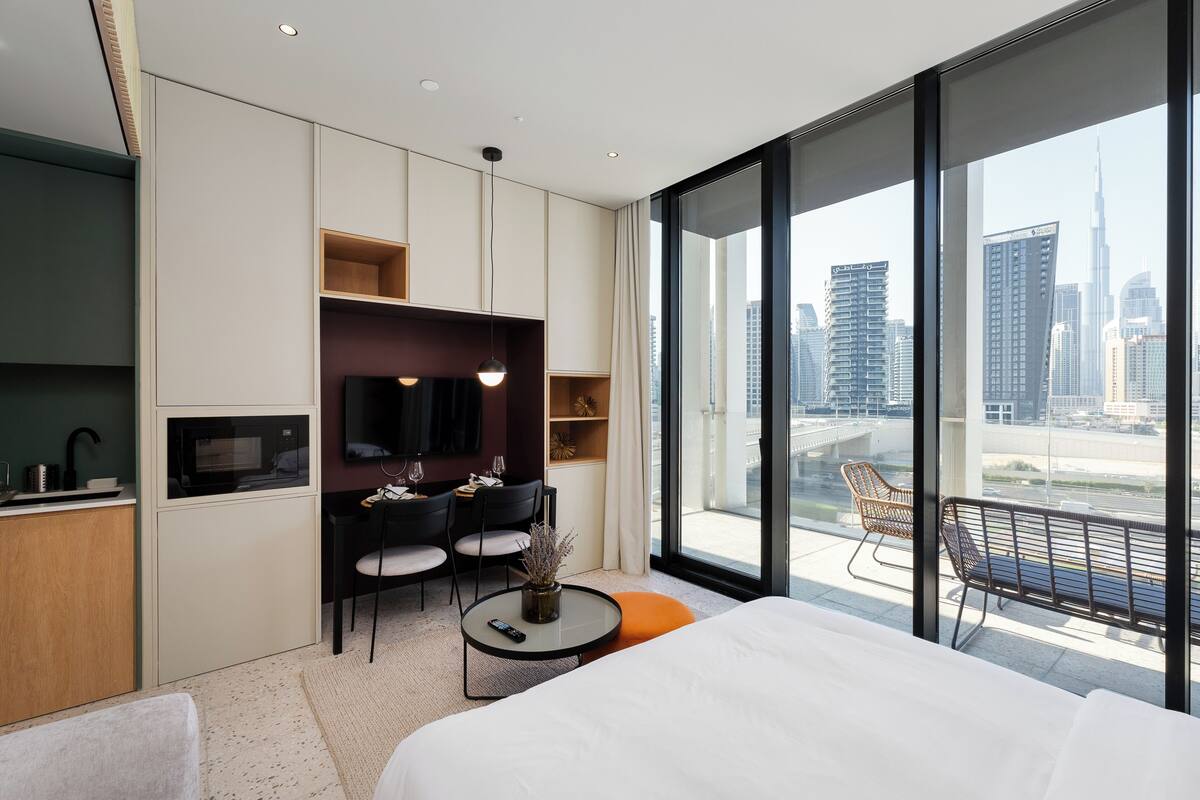 A modern hotel room with a large bed, a dining area with chairs, a small table, and a view of the city skyline through large windows.