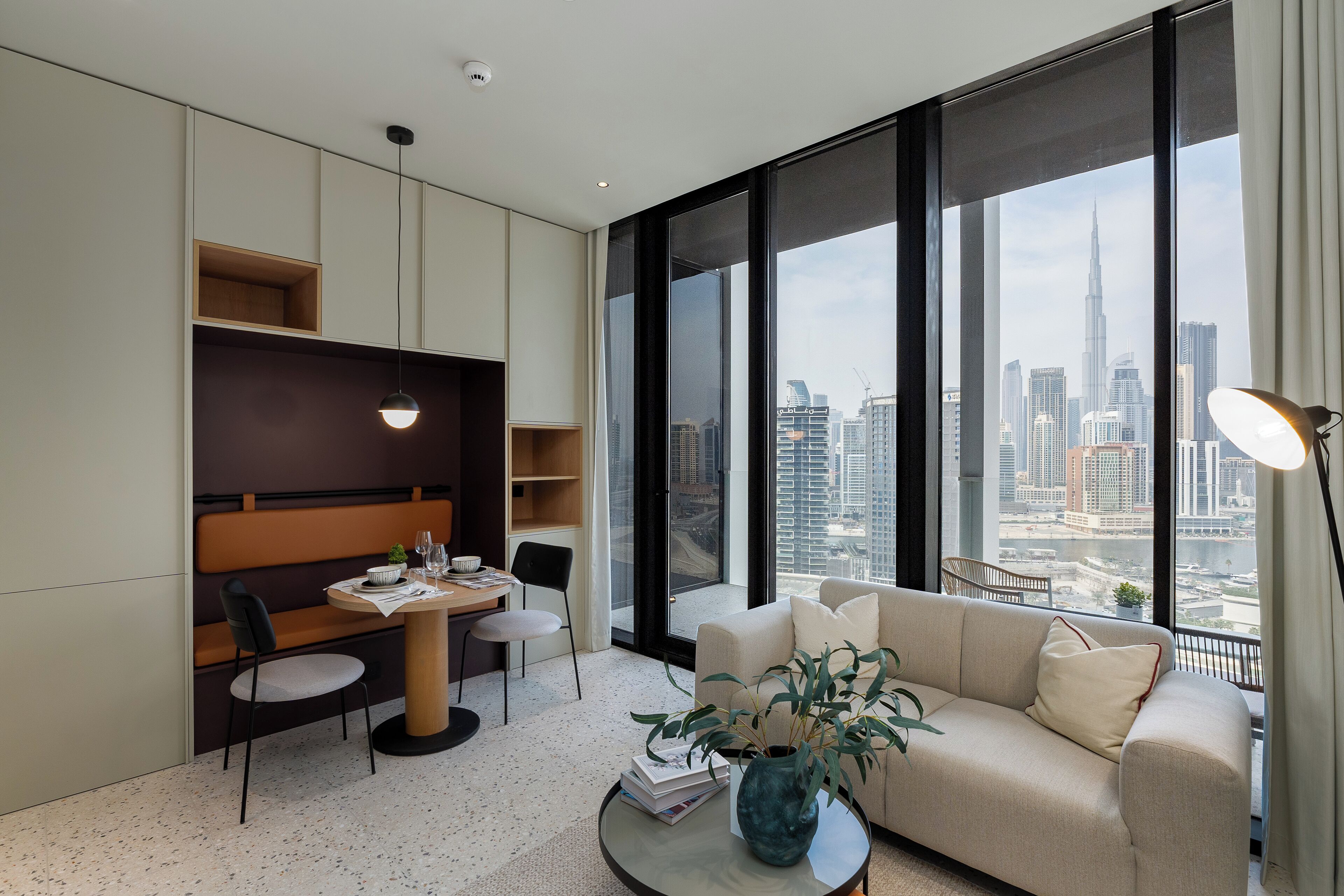 A modern living room with a sofa, dining table, and chairs, large windows with a city view, and a potted plant.