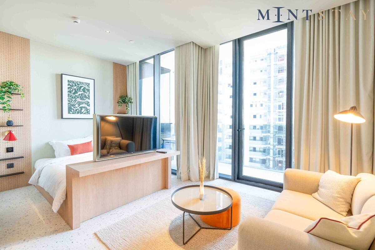 A modern hotel room with a bed, a desk, a sofa, and a large window offering a city view.
