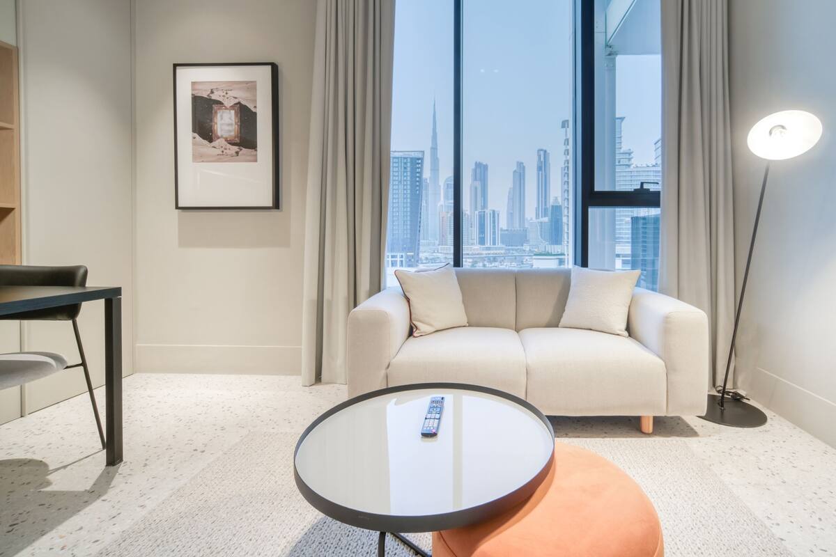 A modern hotel room with a sofa, a round table, a framed picture, and a view of a city skyline through large windows.