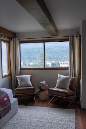 A bedroom with a large window, two chairs, a small round table, and a bed with a white cross on the blanket. - Romantic Swiss Chalet with Mountain Views (Golden)