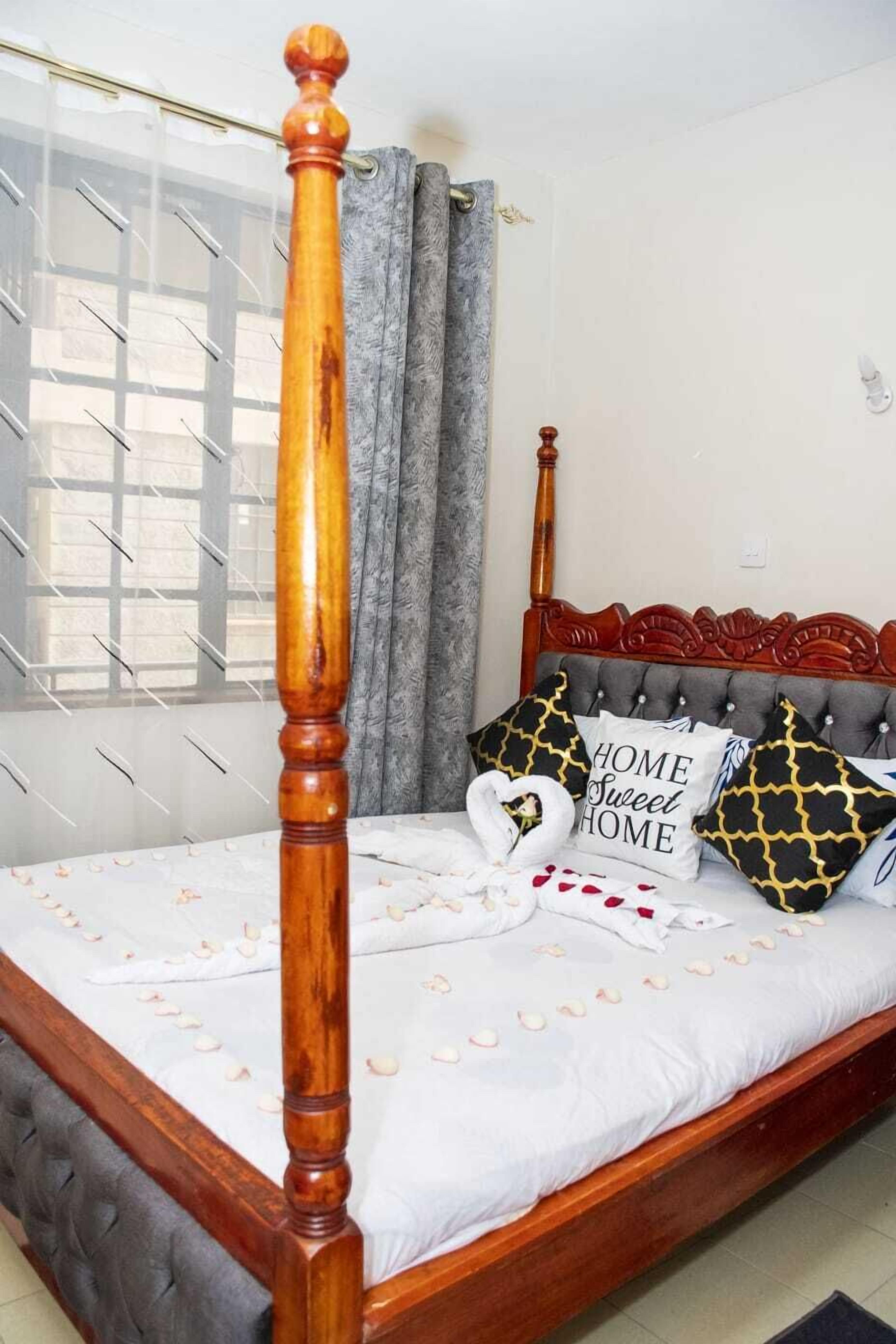 Una cama con dosel y tablero de cabecera de madera, una colcha blanca con pétalos de rosa y almohadones decorativos con texto.