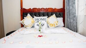 Ein ordentlich bezogenes Bett mit einem floralen Kopfkissen, einem Dekokissen mit dem Schriftzug „HOME“ und einer einzelnen Rose.