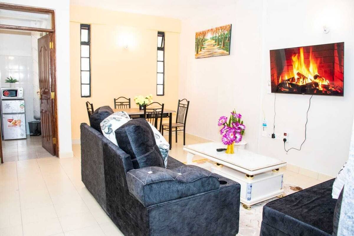 A living room with a sofa, a coffee table with a vase of flowers, a dining area with chairs, and a TV mounted on the wall.