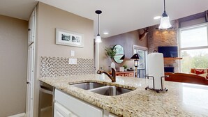 A kitchen with granite countertops, stainless steel sink, pendant lights, a stone fireplace, and a large window. - River City Resorts: TBM 121: Spacious Hill Country Condo at T Bar M Ranch (New Braunfels)