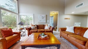 A spacious living room with leather sofas, a wooden coffee table, and a large window offering a view of trees. - River City Resorts: TBM 121: Spacious Hill Country Condo at T Bar M Ranch (New Braunfels)