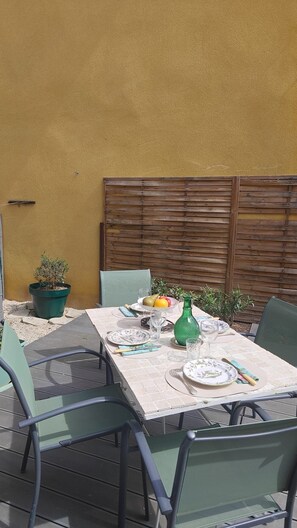 Un espace de restauration en plein air avec une table dressée pour quatre personnes, entourée de chaises, adossé à un mur jaune et bordé d’une clôture en bois.