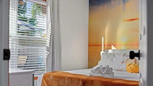 A hotel room with a bed, a window with blinds, and a sunset mural on the wall.