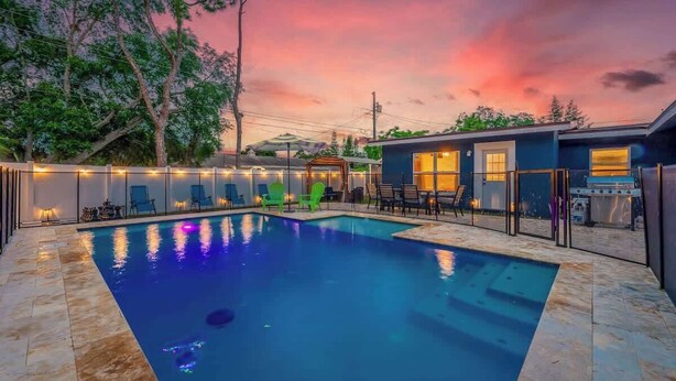 A swimming pool with a stone border, surrounded by lounge chairs and a barbecue area, under a twilight sky.