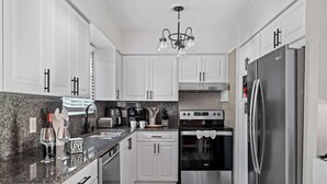 A modern kitchen with white cabinets, granite countertops, stainless steel appliances, and a chandelier.