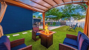 An outdoor patio area with a fire pit, wicker furniture, and a view of a pool and greenery.